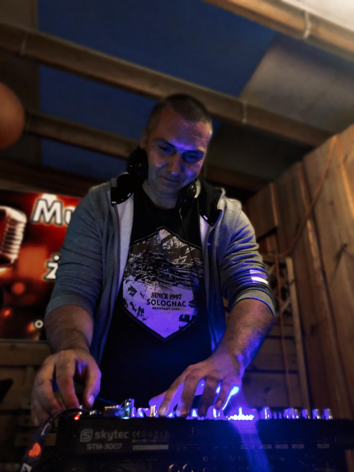 Mężczyzna w słuchawkach, w szarej bluzie z kapturem, obsługuje konsoletę Skytec STM-3007 w fioletowym oświetleniu, w tle częściowo widoczny plakat z mikrofonem.