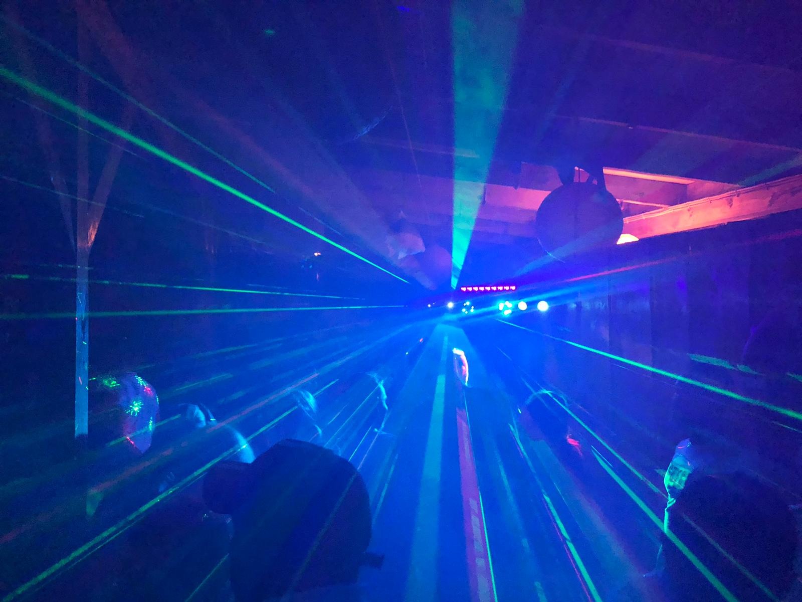Intensywne niebieskie i zielone lasery przecinają ciemne pomieszczenie z widocznymi elementami konstrukcyjnymi sufitu i dyskotekową kulą. Reflektory na suficie tworzą punkt centralny.