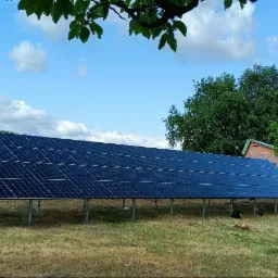Długa instalacja paneli słonecznych na metalowej konstrukcji, umieszczona na zewnątrz, na trawiastym terenie, z widocznym budynkiem w tle i błękitnym niebem z chmurami.