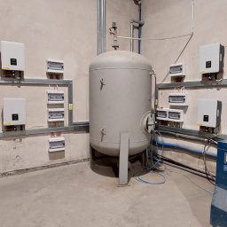Infinite Energy Patryk Wiśniewski - Szafy sterownicze i falowniki zamontowane na ścianie w pomieszczeniu technicznym, widoczny także zbiornik ciśnieniowy i kompresor.
