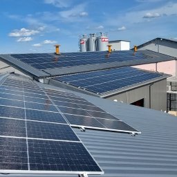Infinite Energy Patryk Wiśniewski - Instalacja paneli słonecznych na dachu budynku przemysłowego, w tle silosy i składowane materiały budowlane.