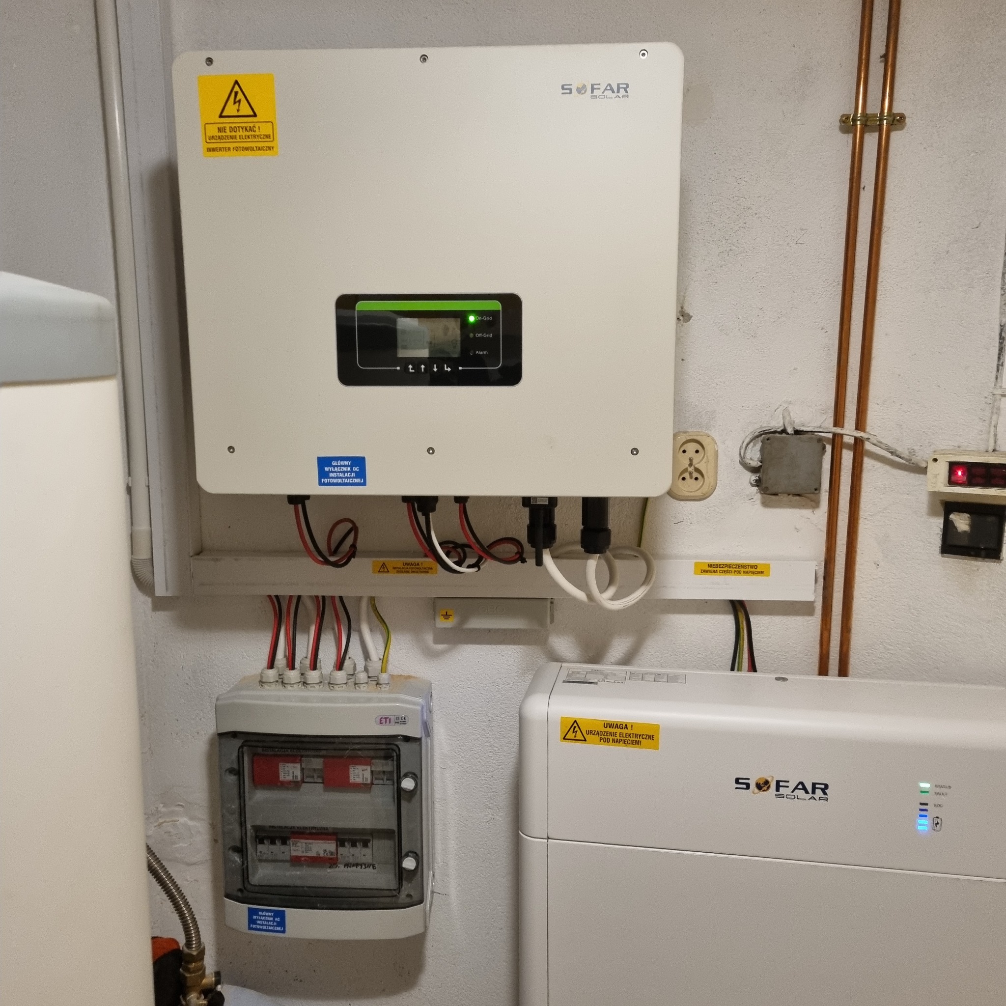 Inwerter SOFAR podłączony do instalacji elektrycznej z zabezpieczeniami, widoczne przewody, tabliczka ostrzegawcza o napięciu i schemat elektryczny w tle. Na ścianie rury miedziane.