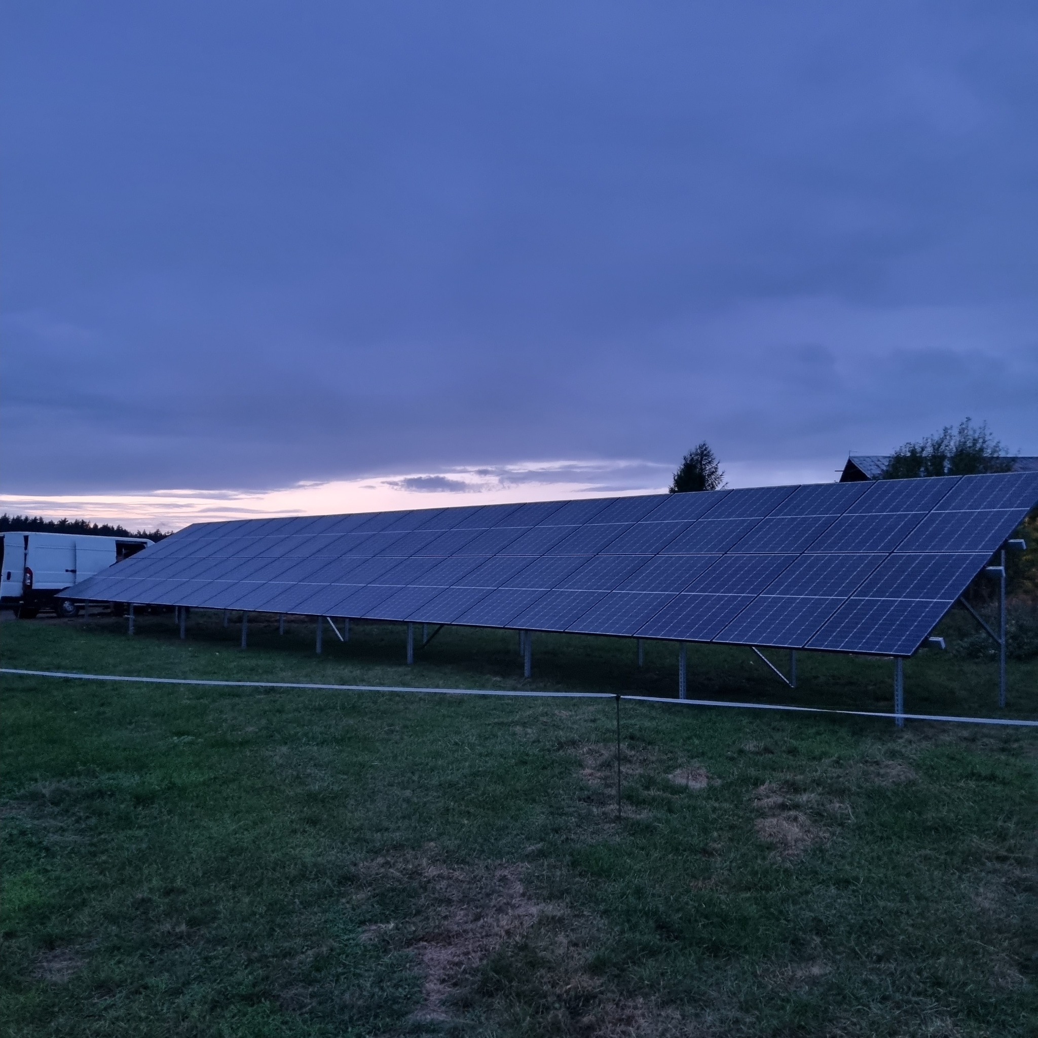 Instalacja fotowoltaiczna na metalowej konstrukcji na trawiastym terenie, z białym busem w tle, sfotografowana o zmierzchu.