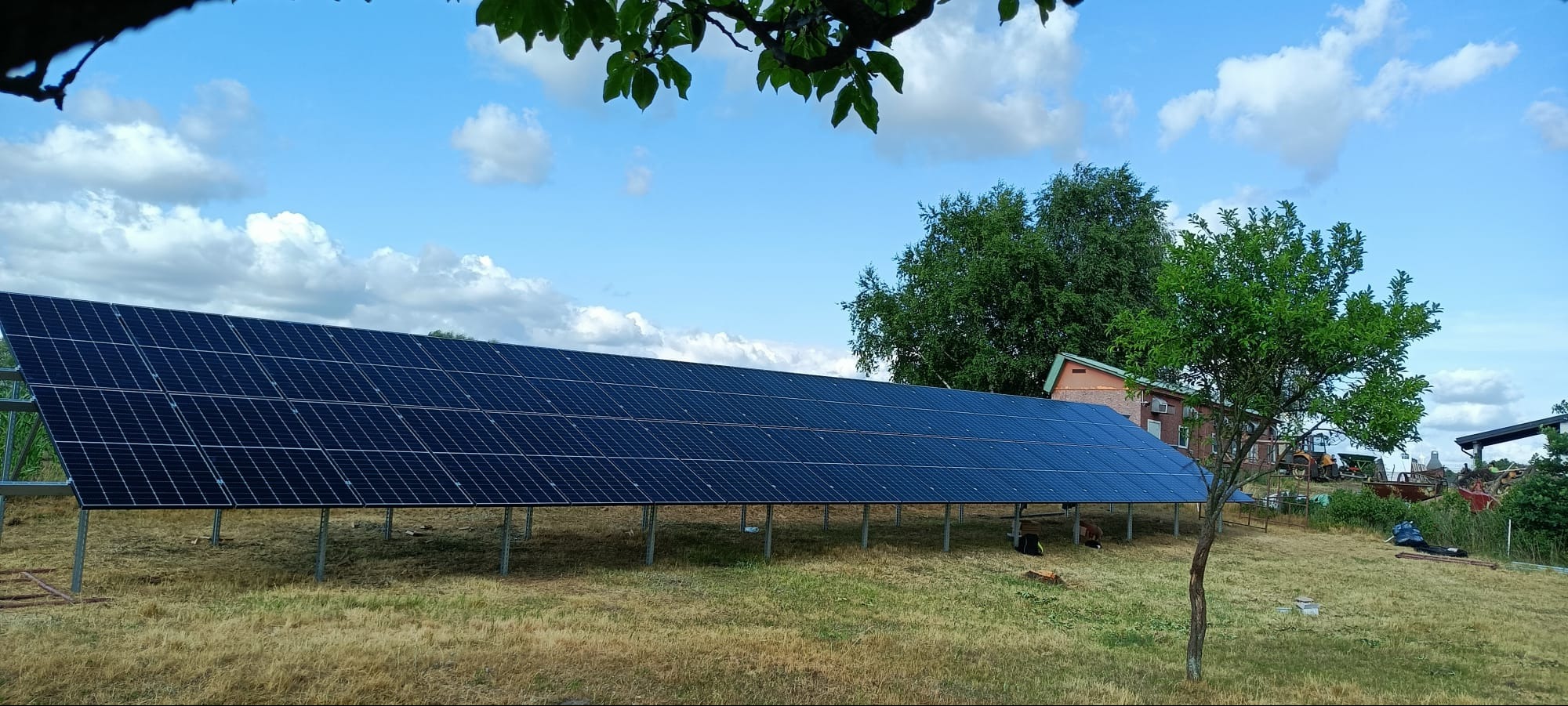 Długa instalacja paneli słonecznych na metalowej konstrukcji, umieszczona na zewnątrz, na trawiastym terenie, z widocznym budynkiem w tle i błękitnym niebem z chmurami.