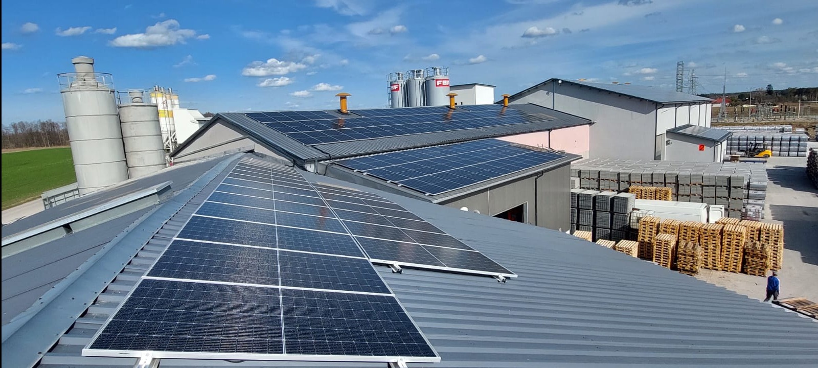 Instalacja paneli słonecznych na dachu budynku przemysłowego, w tle silosy i składowane materiały budowlane.