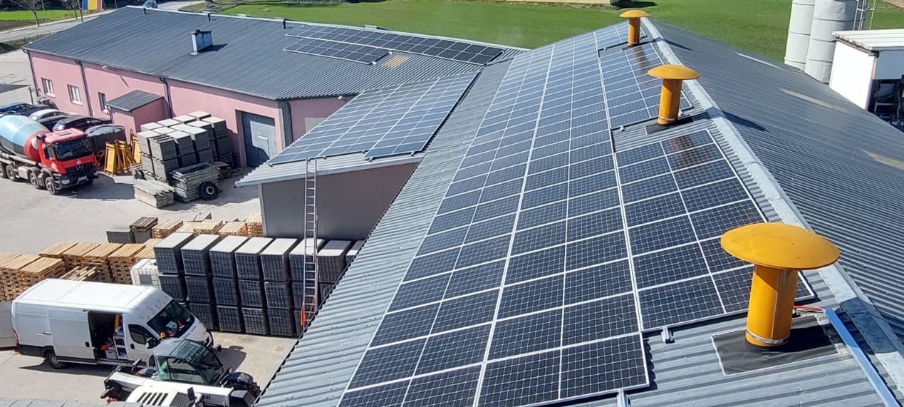 Instalacja fotowoltaiczna na blaszanym dachu budynku przemysłowego z żółtymi kominkami wentylacyjnymi. W tle zaparkowane pojazdy i składowane materiały budowlane.