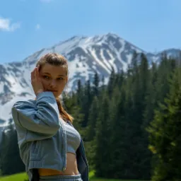 Portret młodej kobiety w sportowym stroju na tle górskiego krajobrazu z lasem i ośnieżonym szczytem.