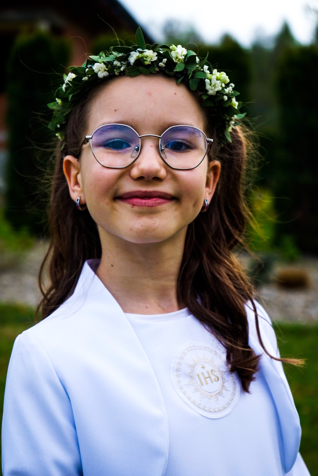 Portret uśmiechniętej dziewczynki w okularach i wianku z białych kwiatów, ubrana w białą albę z symbolem IHS, na tle zieleni.