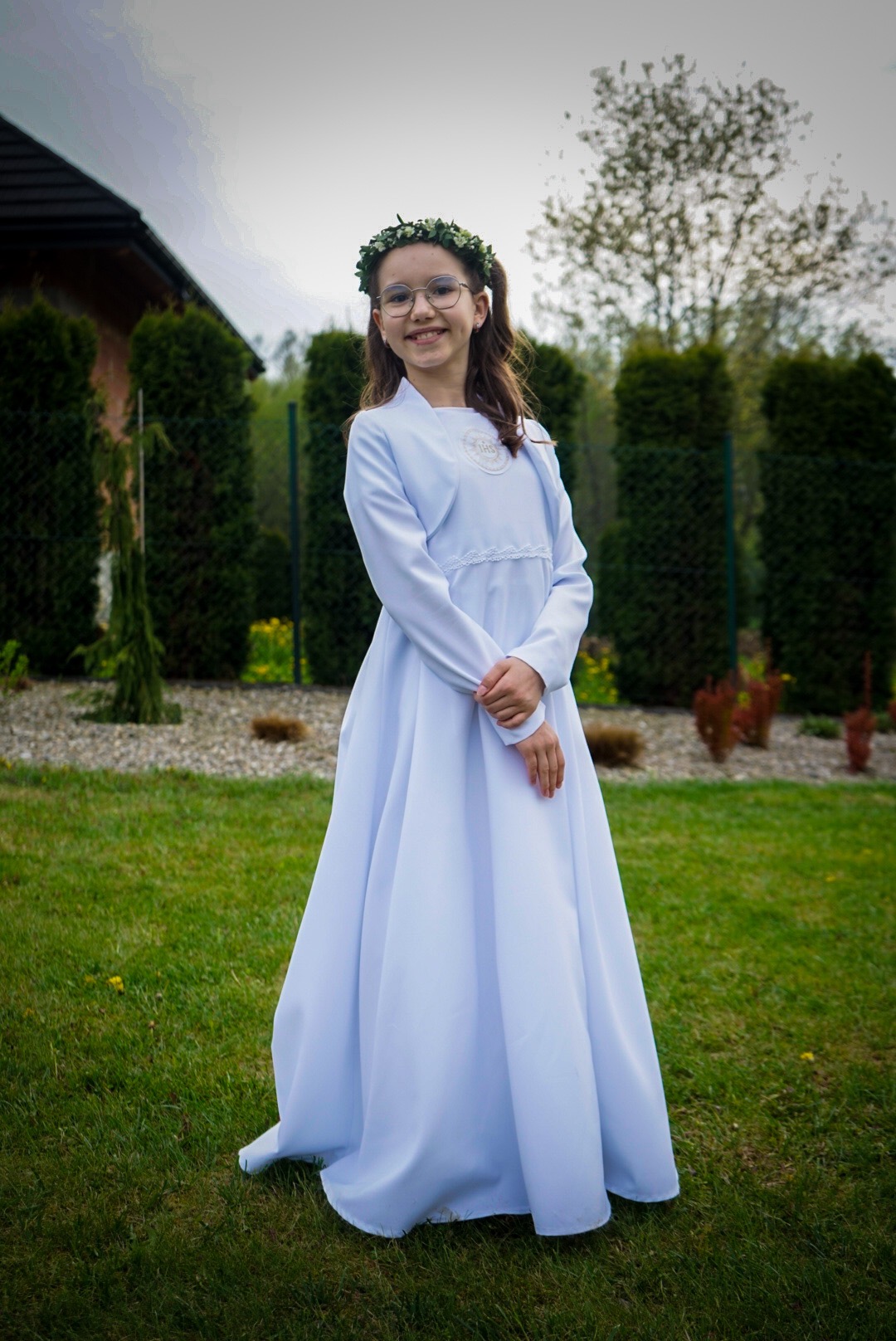 Dziewczynka w okularach i białej albie komunijnej z wiankiem na głowie, pozująca na trawniku przed domem.