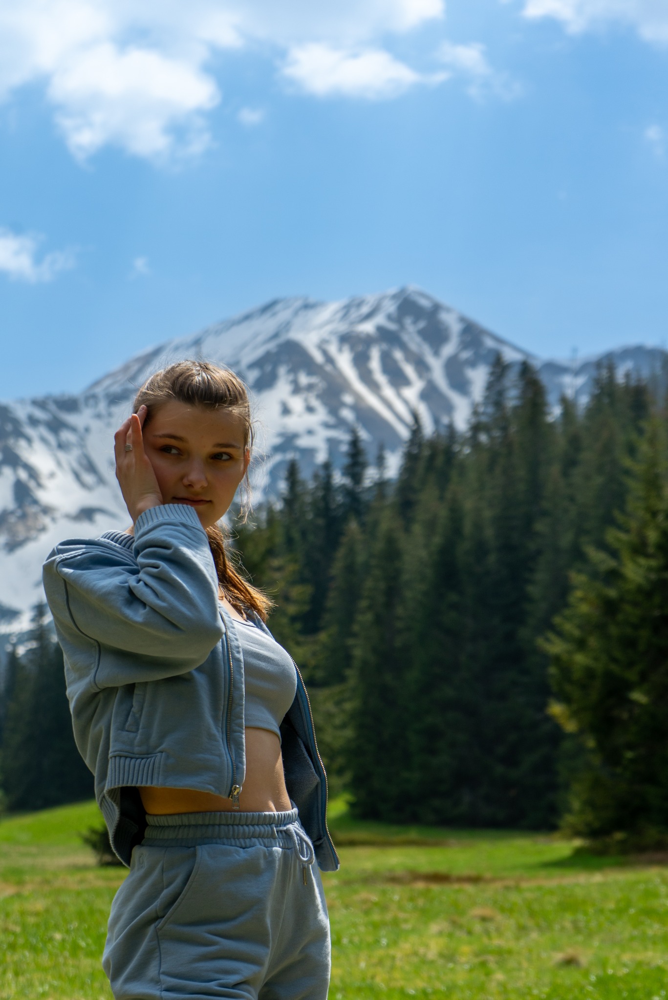Portret młodej kobiety w sportowym stroju na tle górskiego krajobrazu z lasem i ośnieżonym szczytem.
