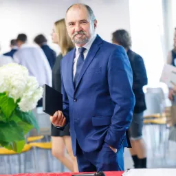 Mężczyzna w niebieskim garniturze trzyma czarne pudełko na tle sali z publicznością i bukietem białych kwiat&oacute;w.