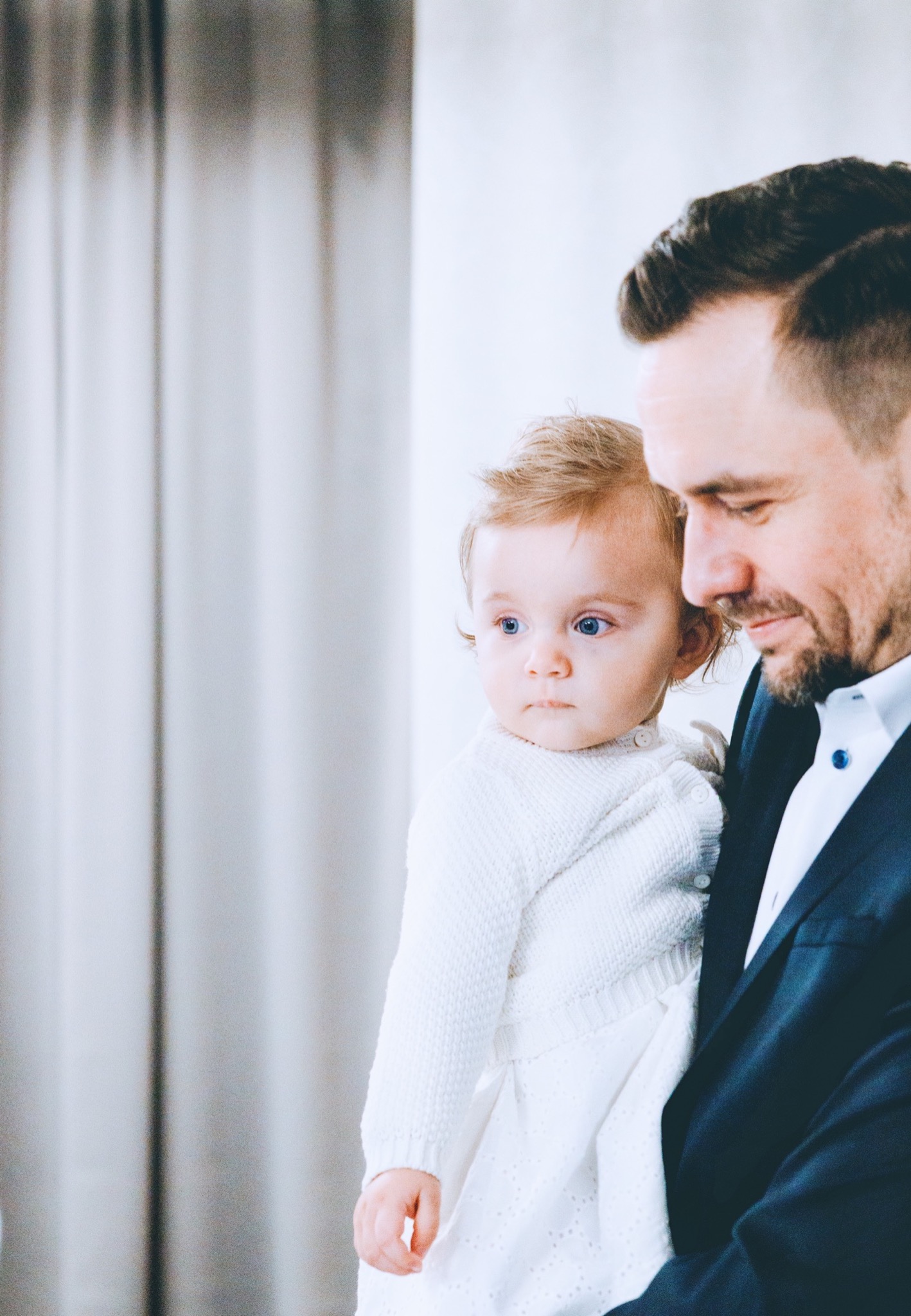 Ojciec w granatowym garniturze trzyma na rękach małą dziewczynkę w białej sukience, oboje patrzą w lewo, neutralne tło.