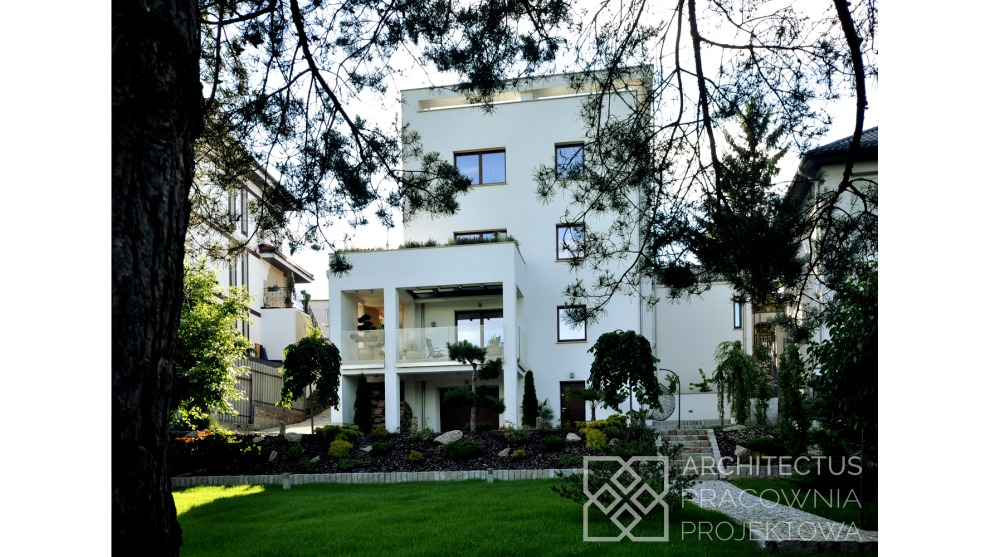 Nowoczesny, trzypiętrowy dom jednorodzinny z płaskim dachem, białą elewacją i balkonem z przeszkloną balustradą, otoczony zielenią i drzewami, częściowo przesłonięty gałęziami drzewa na pierwszym...