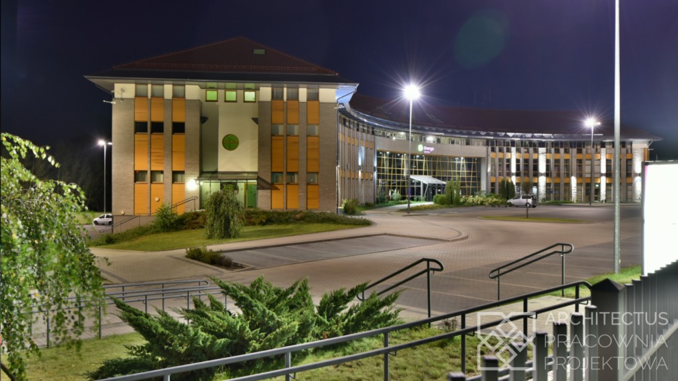 Oświetlony nocą budynek biurowy z elementami drewnianymi i szklaną fasadą, widoczny z perspektywy schodów z metalową barierką, zieleń w otoczeniu i logo firmy na budynku.