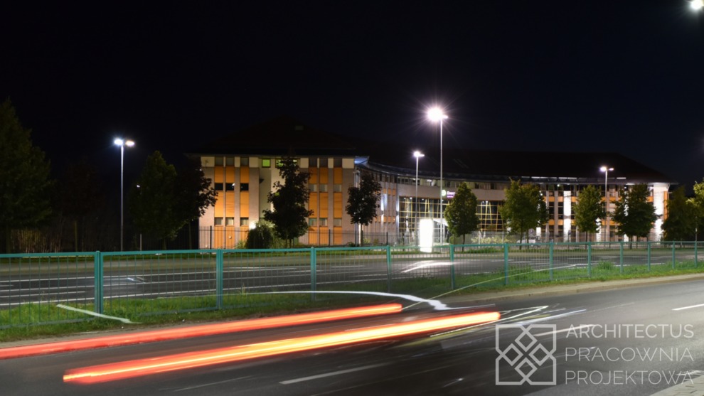 Nocne ujęcie budynku usługowego z pomarańczowymi panelami, widoczne smugi świateł samochodowych, oświetlenie uliczne i logo firmy projektowej w prawym dolnym rogu.