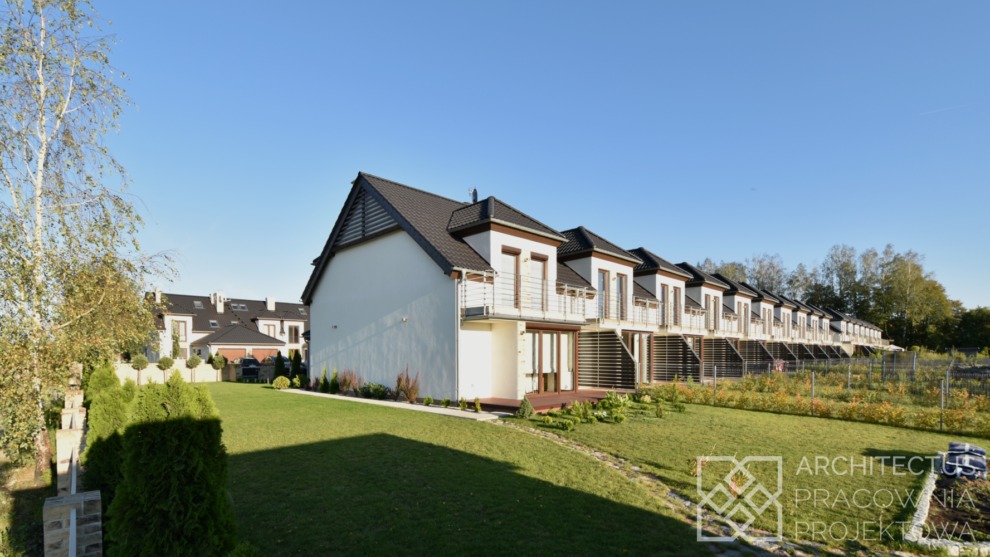 Szereg identycznych domów jednorodzinnych z balkonami, w jasnej kolorystyce, z ciemnymi dachami, otoczonych zielonym trawnikiem i niską roślinnością.