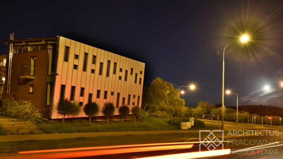 Nowoczesny budynek usługowy z elewacją z paneli w nocnym ujęciu, widoczne smugi świateł samochodowych i logo pracowni projektowej w prawym dolnym rogu.
