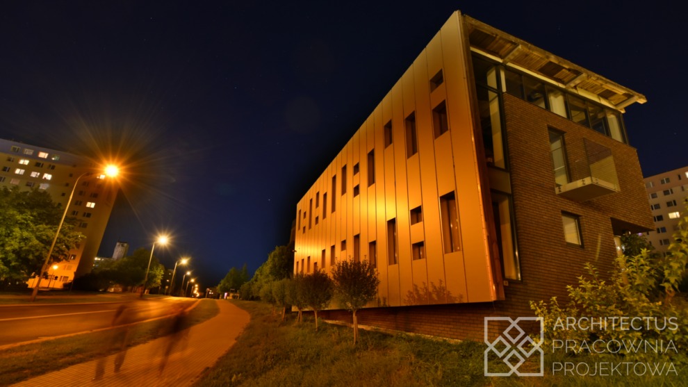 Nowoczesny budynek usługowy z elewacją z paneli w ciepłym oświetleniu nocnym, widoczny chodnik i sylwetki przechodniów, logo pracowni projektowej w prawym dolnym rogu.