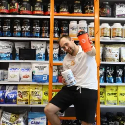 Mężczyzna na drabinie w sklepie z suplementami diety, trzymający pomarańczowy shaker i opakowanie białka jajecznego, p&oacute;łki wypełnione r&oacute;żnymi odżywkami i suplementami dla sportowc&oacute;w w tle.