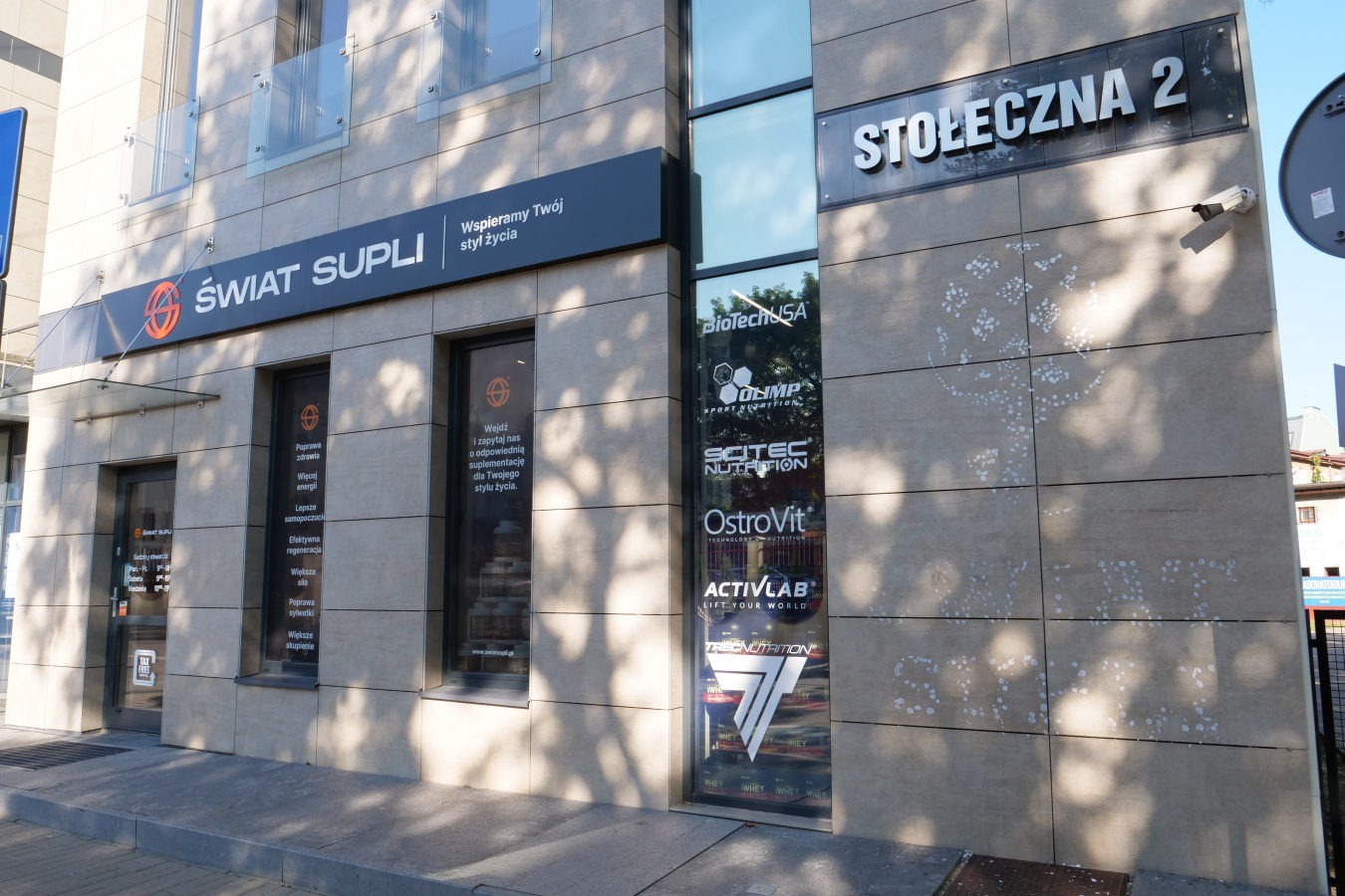 Fasada budynku sklepu z suplementami diety 'Świat Supli' z witrynami oklejonymi reklamami marek suplementów, adres 'Stołeczna 2' na tabliczce.
