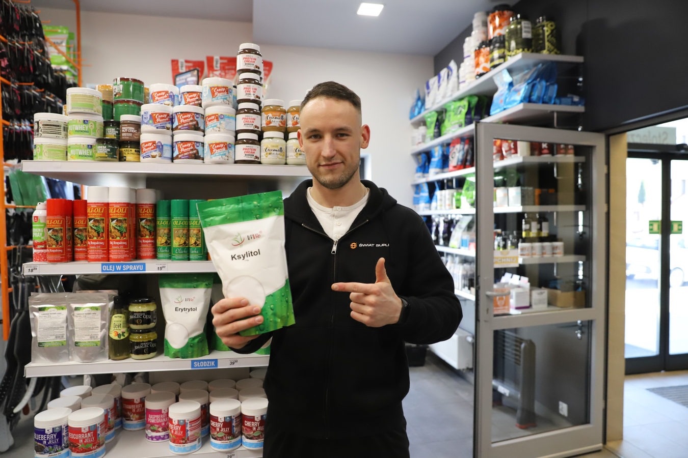 Mężczyzna w czarnej bluzie z logo 'Świat Supli' prezentuje opakowanie ksylitolu marki Vivio na tle półek z produktami spożywczymi i suplementami diety.