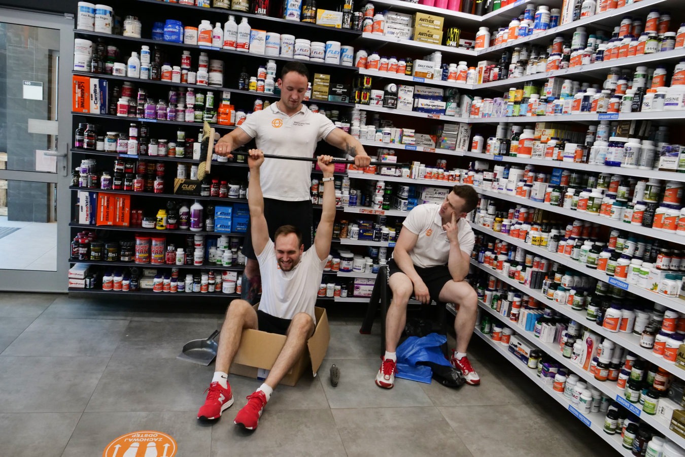 Trzech mężczyzn w sklepie z suplementami diety: jeden siedzi w kartonie unosząc sztangę, drugi stoi nad nim trzymając sztangę, trzeci siedzi na krześle zamyślony, na podłodze naklejka z napisem...