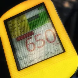 Miernik grubości lakieru samochodowego z żółtą obudową, wyświetlający wynik 650 um, z informacją o pomiarze na szpachli i aluminium, na tle karoserii pojazdu.