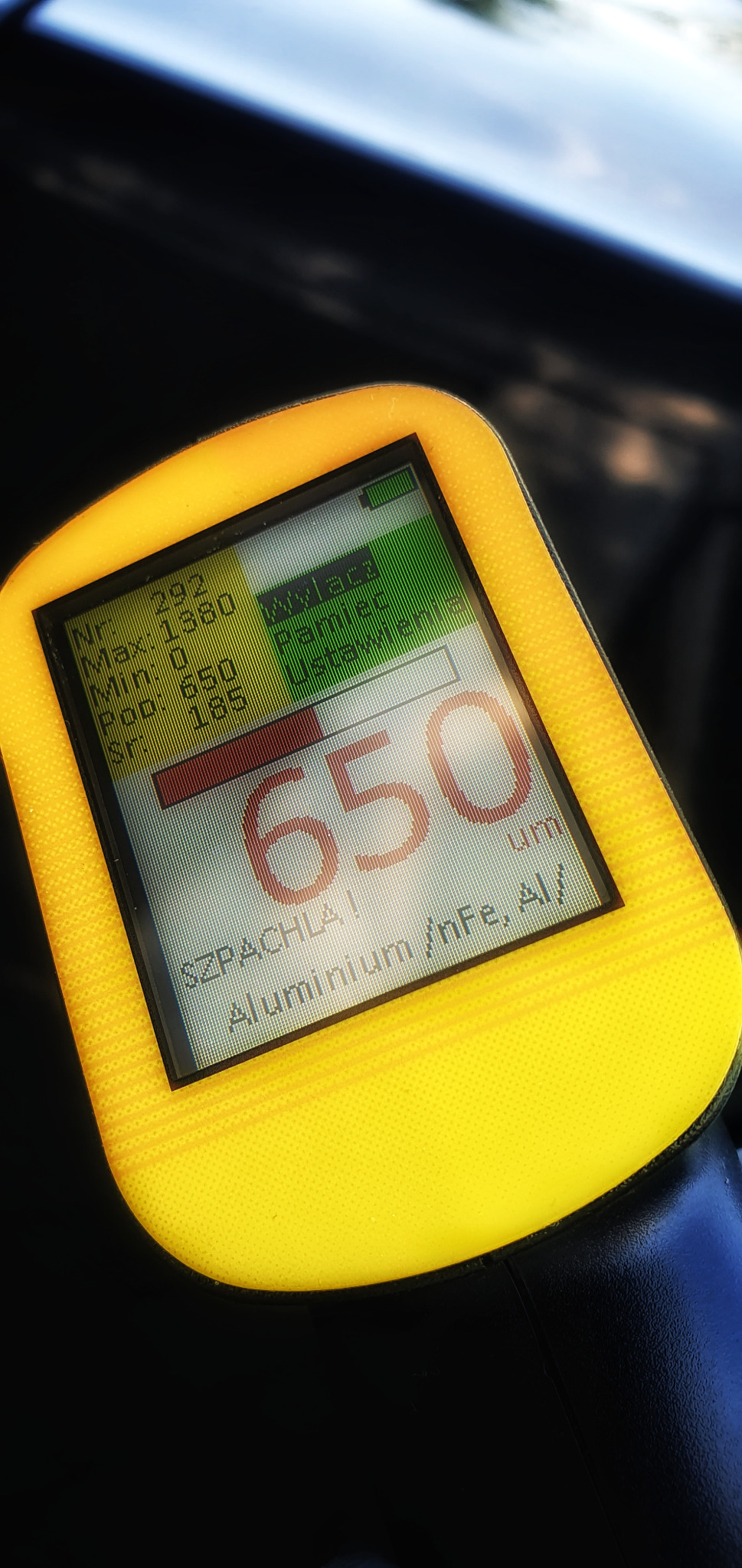 Miernik grubości lakieru samochodowego z żółtą obudową, wyświetlający wynik 650 um, z informacją o pomiarze na szpachli i aluminium, na tle karoserii pojazdu.