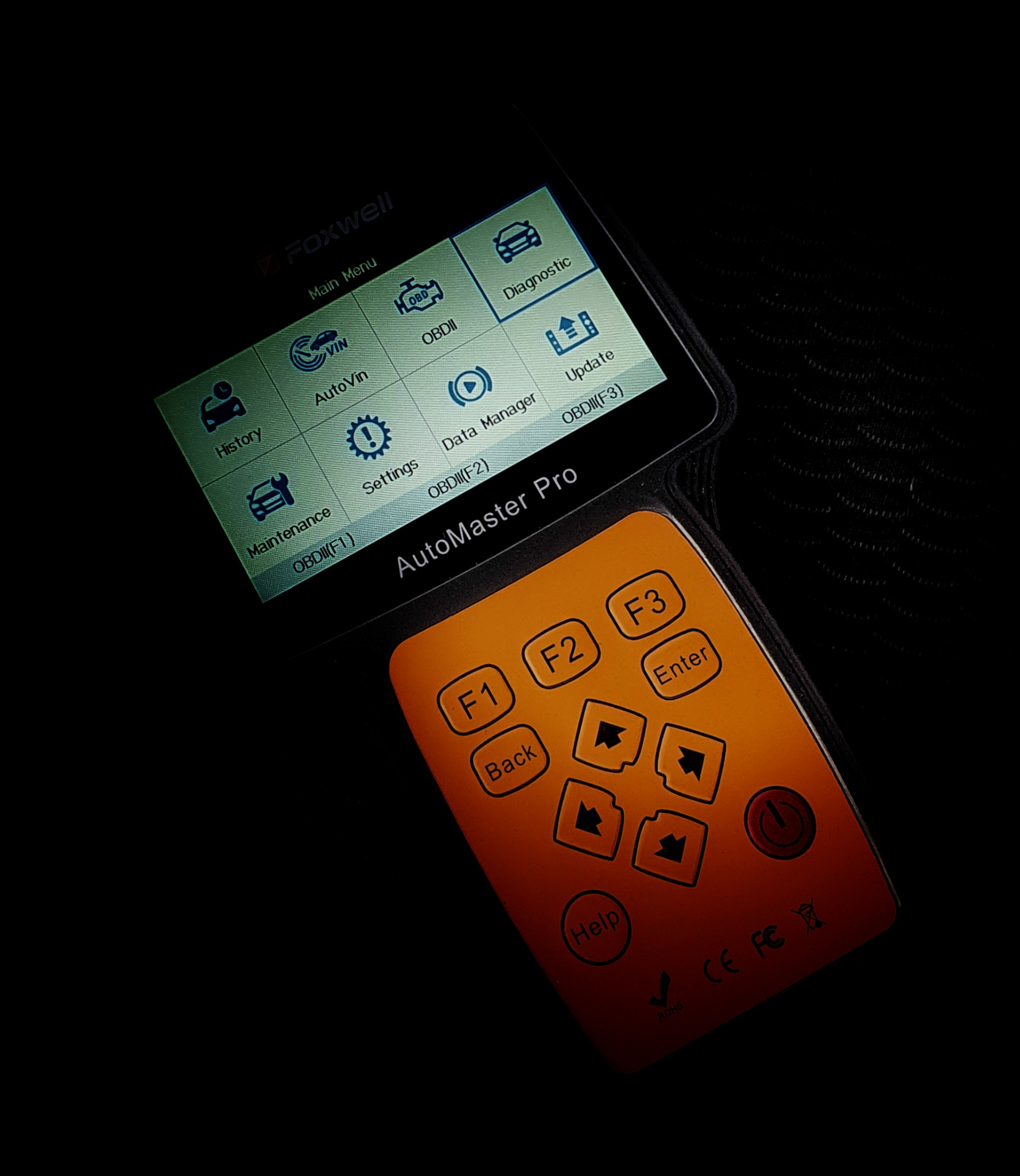 Urządzenie diagnostyczne Foxwell AutoMaster Pro z podświetlonym ekranem menu głównego i pomarańczowym panelem sterowania, leżące na ciemnym tle.