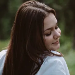 Portret młodej kobiety z długimi, ciemnymi włosami, odwróconej profilem w lewo, z zamkniętymi oczami i delikatnym uśmiechem, ubrana w białą koszulę na tle zieleni.