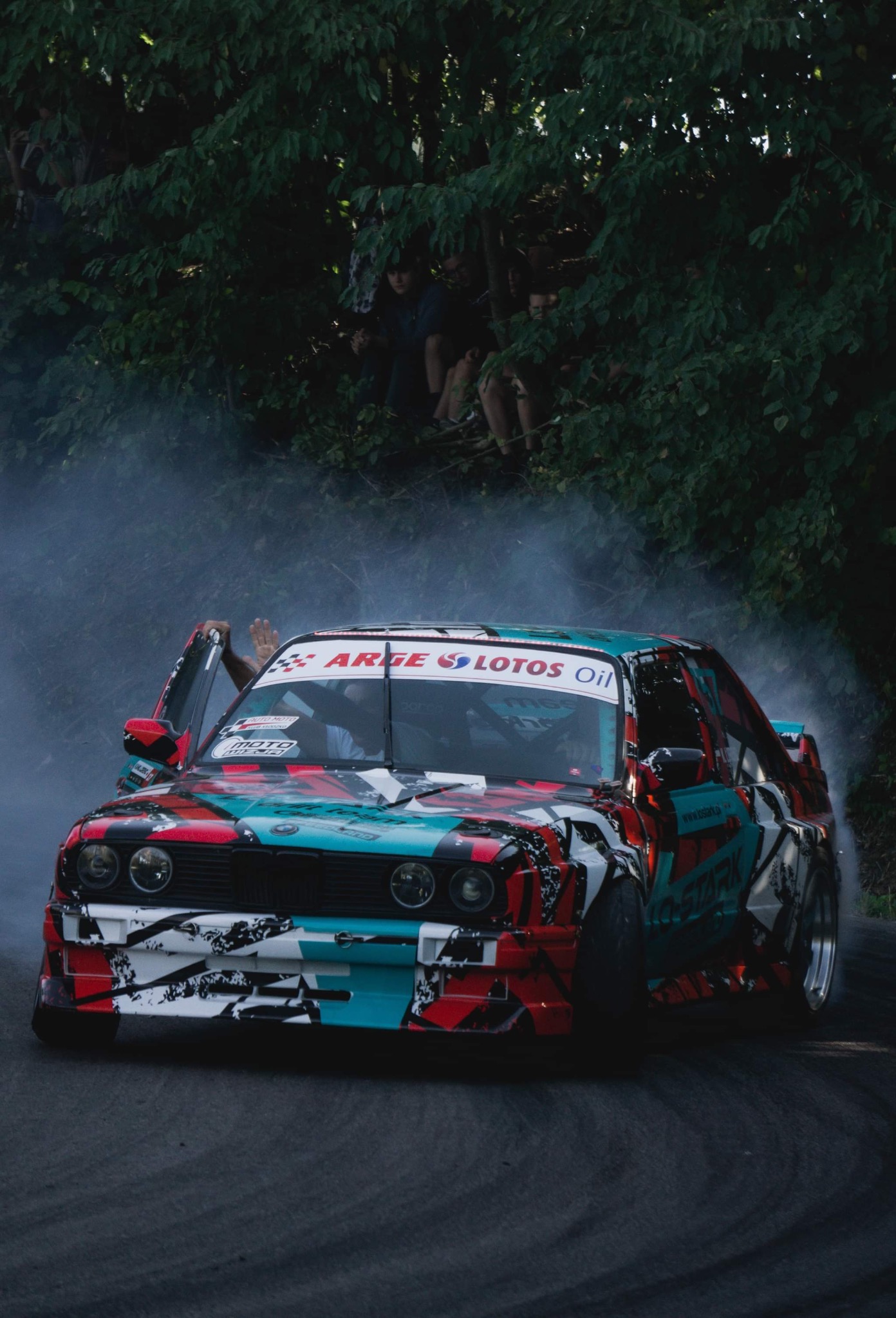 Driftingowy samochód BMW E30 z otwartymi drzwiami kierowcy, oklejony w barwy czerwono-biało-turkusowe, podczas pokazu na torze; w tle obserwujący widzowie.