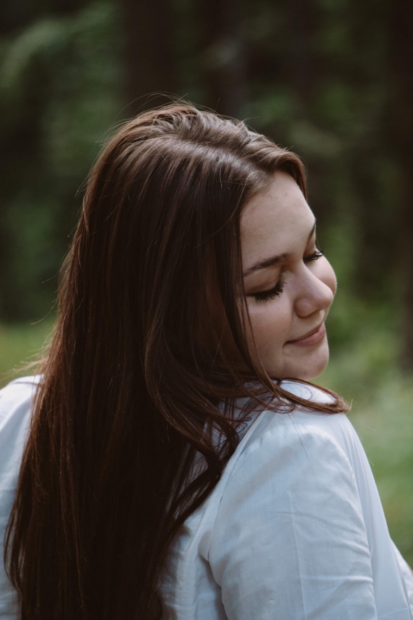 Portret młodej kobiety z długimi, ciemnymi włosami, odwróconej profilem w lewo, z zamkniętymi oczami i delikatnym uśmiechem, ubrana w białą koszulę na tle zieleni.