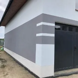 Dwukolorowa elewacja budynku z szarą sekcją środkową i białymi akcentami w narożniku, grafitowa brama garażowa, widoczny fragment rynny i otoczenie w trakcie prac wykończeniowych.