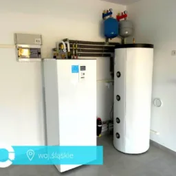 Biała instalacja grzewcza z zbiornikiem akumulacyjnym i naczyniami przeponowymi w jasnym pomieszczeniu z szarą podłogą.