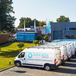Passive Instal Sp. z o.o. - Marcin Gorajek - Flota białych samochodów dostawczych firmy Passive Instal zaparkowana przed nowoczesnym budynkiem biurowym tej samej firmy, na tle zielonego trawnika i drzew.