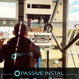 Passive Instal Sp. z o.o. - Marcin Gorajek - Instalator w bluzie z logo firmy Passive Instal podłącza inwerter Growatt do instalacji fotowoltaicznej na zewnątrz budynku.