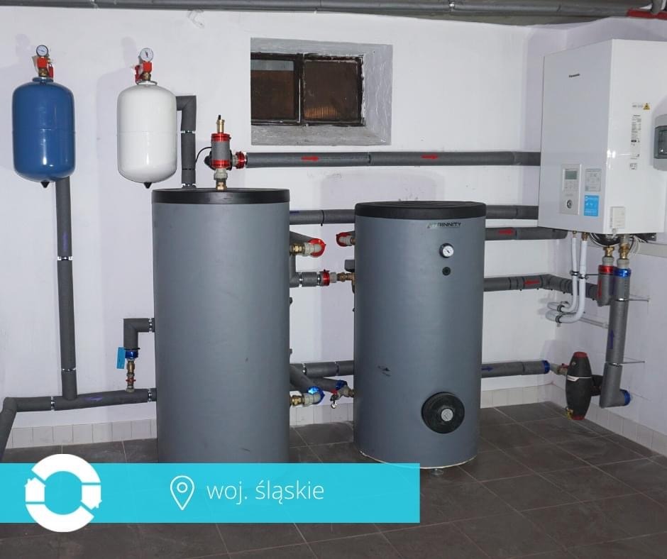 Instalacja dwóch szarych zbiorników buforowych z osprzętem w kotłowni w Częstochowie, z widocznymi rurami w izolacji i białym piecem wiszącym na ścianie.
