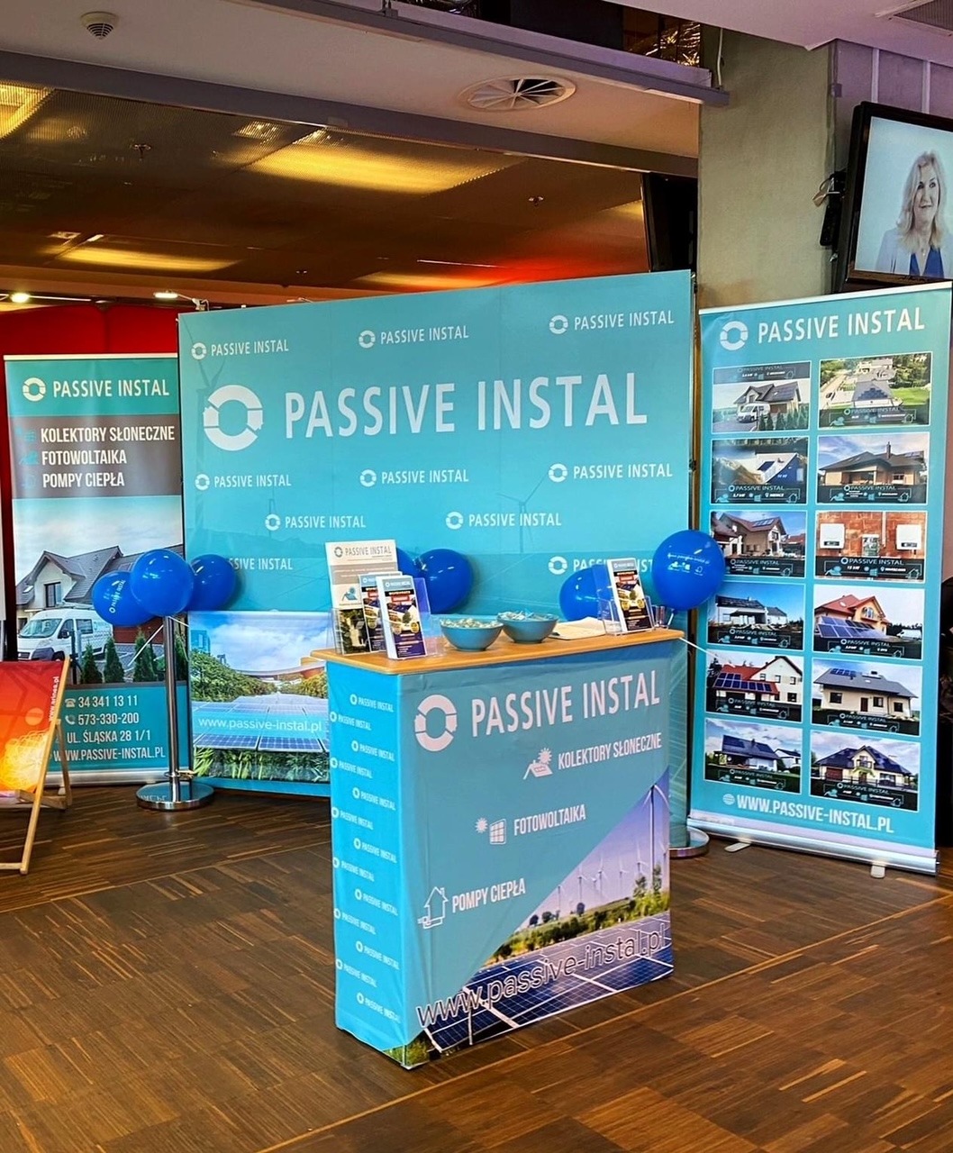Stoisko targowe firmy Passive Instal z banerami reklamowymi prezentującymi usługi: kolektory słoneczne, fotowoltaika i pompy ciepła, udekorowane niebieskimi balonami.