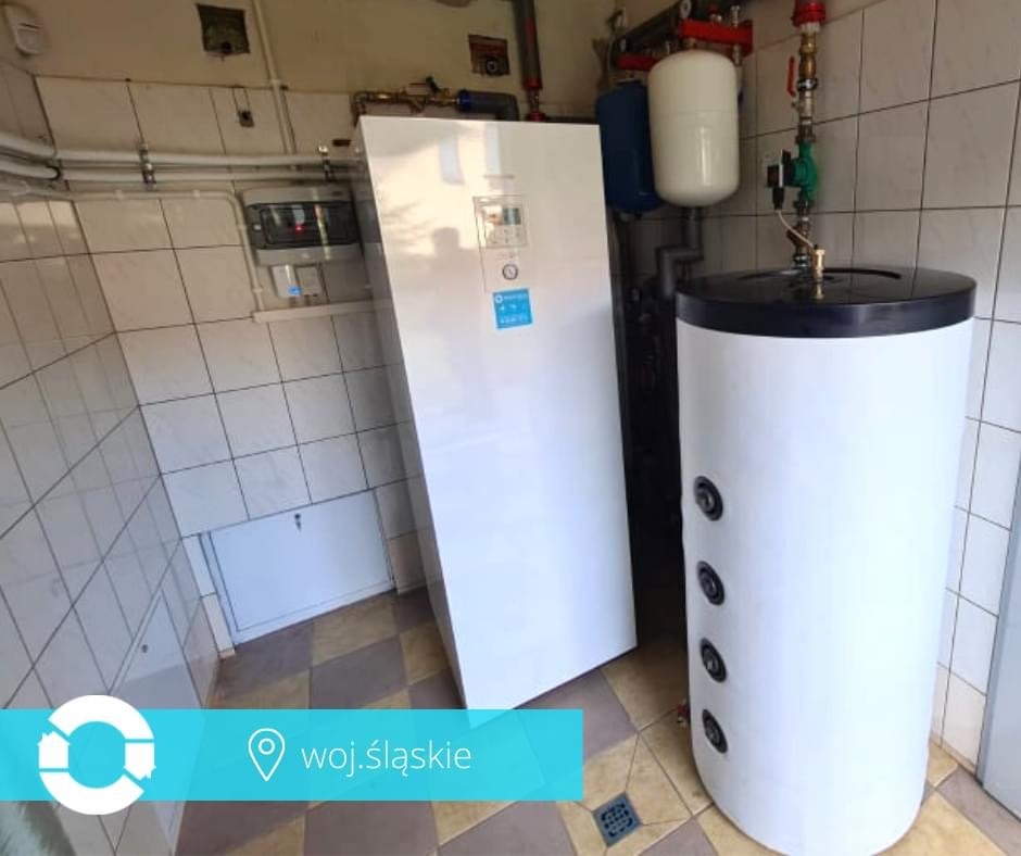 Biała pompa ciepła wisząca na ścianie w pomieszczeniu z biało-szarą glazurą i stojący obok biały zbiornik na wodę z czarnym wieczkiem.