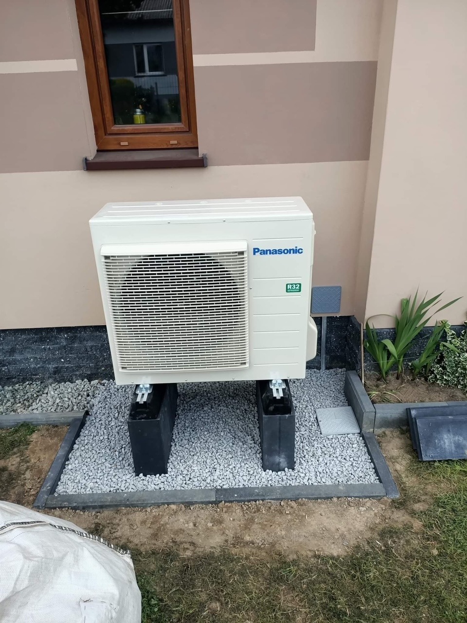 Biała jednostka zewnętrzna klimatyzacji Panasonic R32, zamontowana na czarnych podstawach na tle ściany budynku z oknem i ozdobnym pasem.