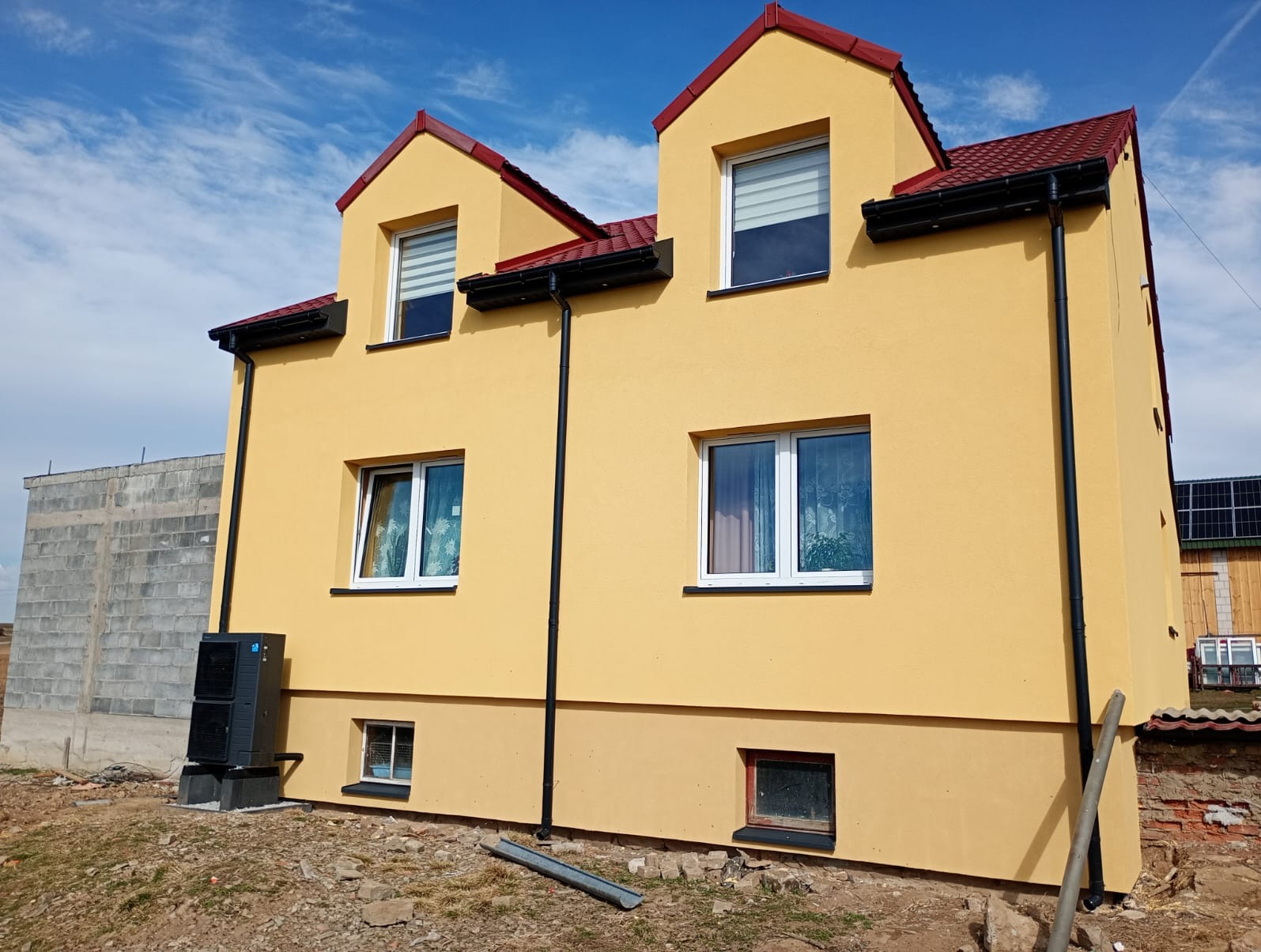 Żółty dom po dociepleniu z widoczną pompą ciepła i panelami słonecznymi na sąsiednim budynku, czarne rynny, czerwony dach, błękitne niebo.