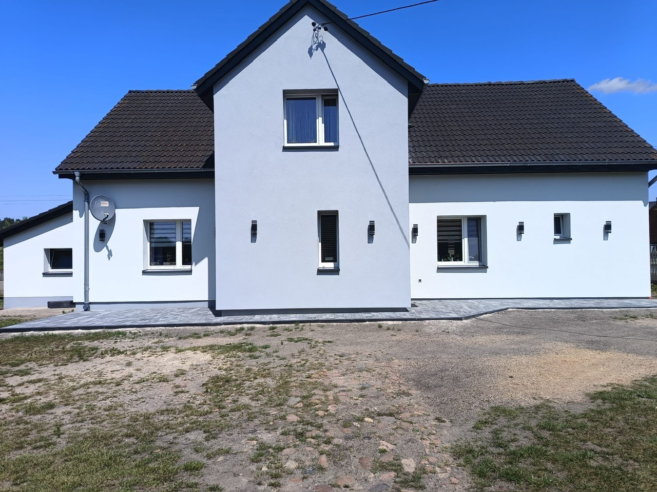 Dom po ociepleniu elewacji styropianem grafitowym i położeniu struktury tynku. Widoczne szare ściany, ciemny dach i kostka brukowa wokół budynku.