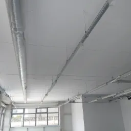 Wnętrze biurowe po remoncie, świeżo pomalowane ściany na biało, widoczne instalacje pod sufitem, rury wentylacyjne w srebrnej izolacji, metalowe korytka kablowe, duże okna z widokiem na zewnątrz...