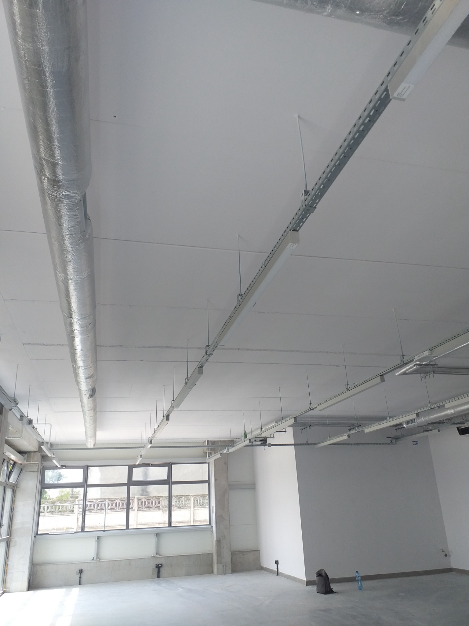 Wnętrze biurowe po remoncie, świeżo pomalowane ściany na biało, widoczne instalacje pod sufitem, rury wentylacyjne w srebrnej izolacji, metalowe korytka kablowe, duże okna z widokiem na zewnątrz...