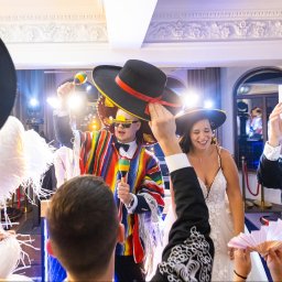 Michał Zaczek - Weselna zabawa z meksykańskim motywem: para młoda w otoczeniu gości w sombrerach i ponczach, mężczyzna gra na marakasach, w tle bogato zdobiona sala.