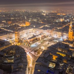 Michał Zaczek - Nocny widok z lotu ptaka na ośnieżony Rynek Główny w Krakowie, oświetlony budynki Sukiennic, Kościół Mariacki i okoliczne kamienice, z widoczną choinką na placu i światłami miasta w tle.