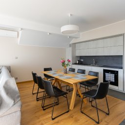 Michał Zaczek - Jasne wnętrze mieszkania w Bielsko-Białej z drewnianym stołem, czarnymi krzesłami, szarą sofą i minimalistyczną kuchnią z szarymi szafkami oraz kwiatami w wazonie na stole.