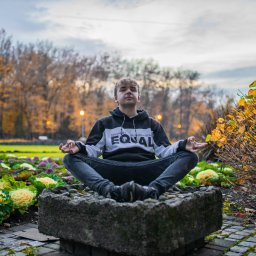 Michał Zaczek - Młody mężczyzna w bluzie z kapturem siedzi w pozycji lotosu na kamiennym postumencie w parku, z rabatami kwiatowymi i jesiennymi drzewami w tle.