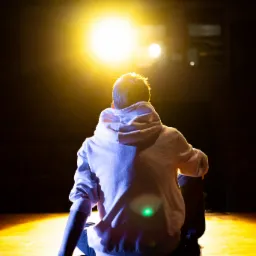 Osoba siedząca na podłodze sceny, oświetlona reflektorem od tyłu, w białej bluzie z kapturem.