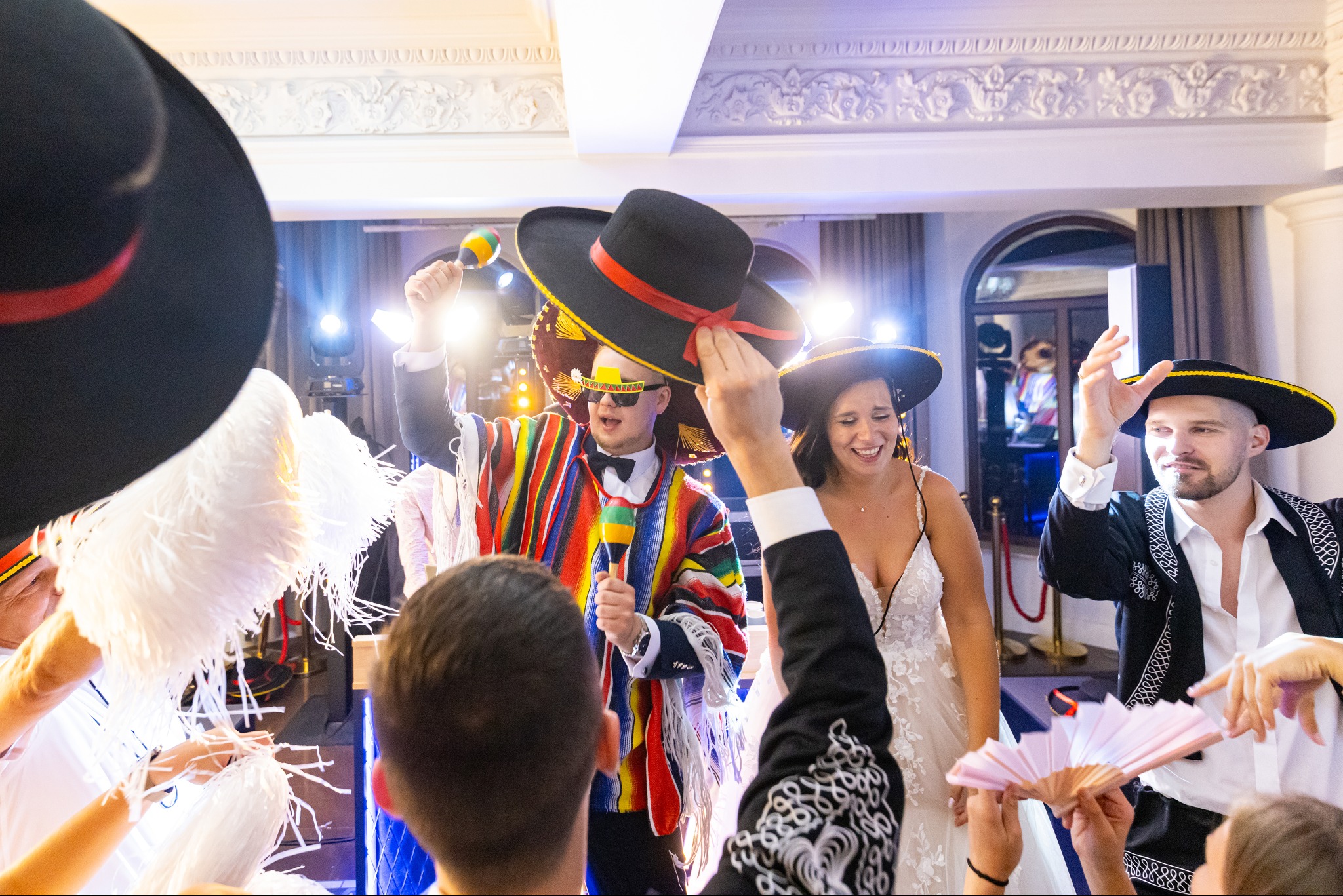 Weselna zabawa z meksykańskim motywem: para młoda w otoczeniu gości w sombrerach i ponczach, mężczyzna gra na marakasach, w tle bogato zdobiona sala.